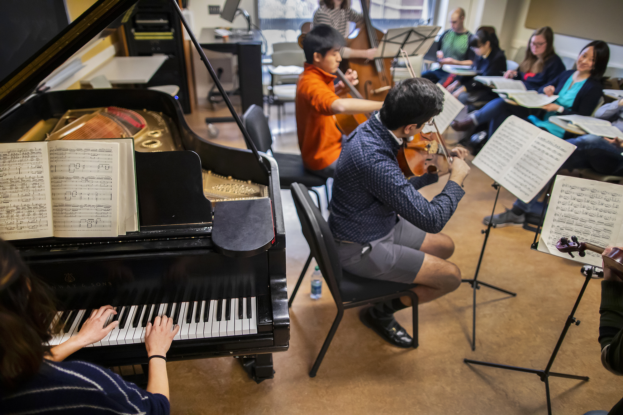 A view that shows a student's hands playing the piano and other students playing the viola, cello and bass in front of class.