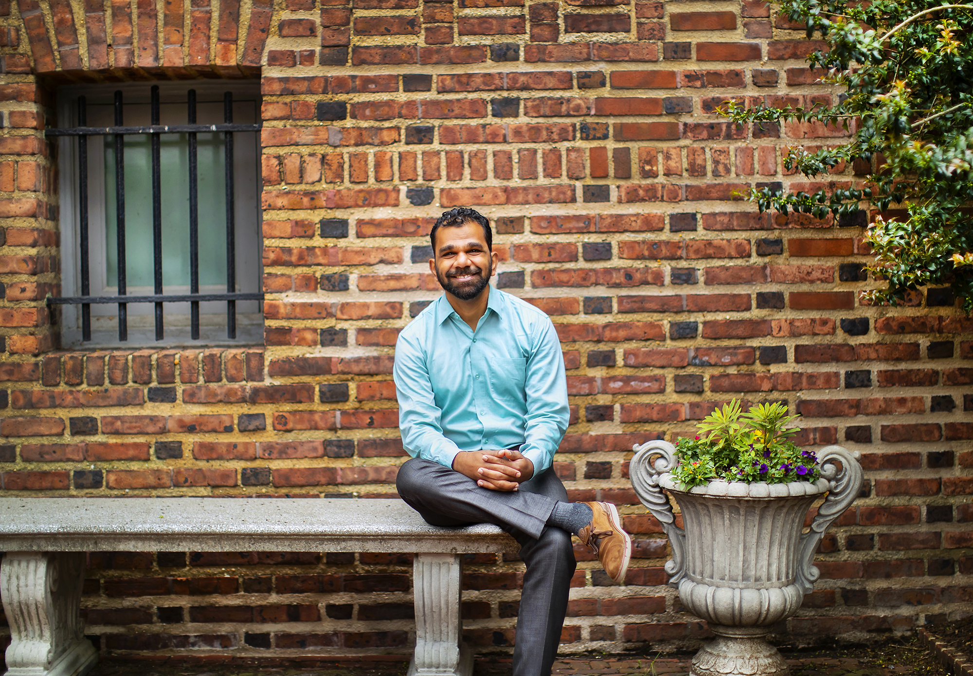 A person in a blue shirt and gray pants sitting on a stone bench in front of a brick building.