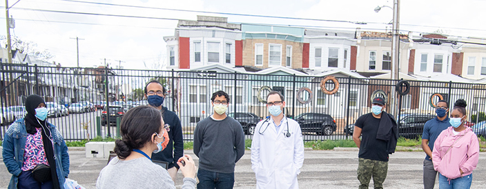 Health care worker stands outside wearing a mask addressing several people in the community who are also wearing a mask and social distancing.
