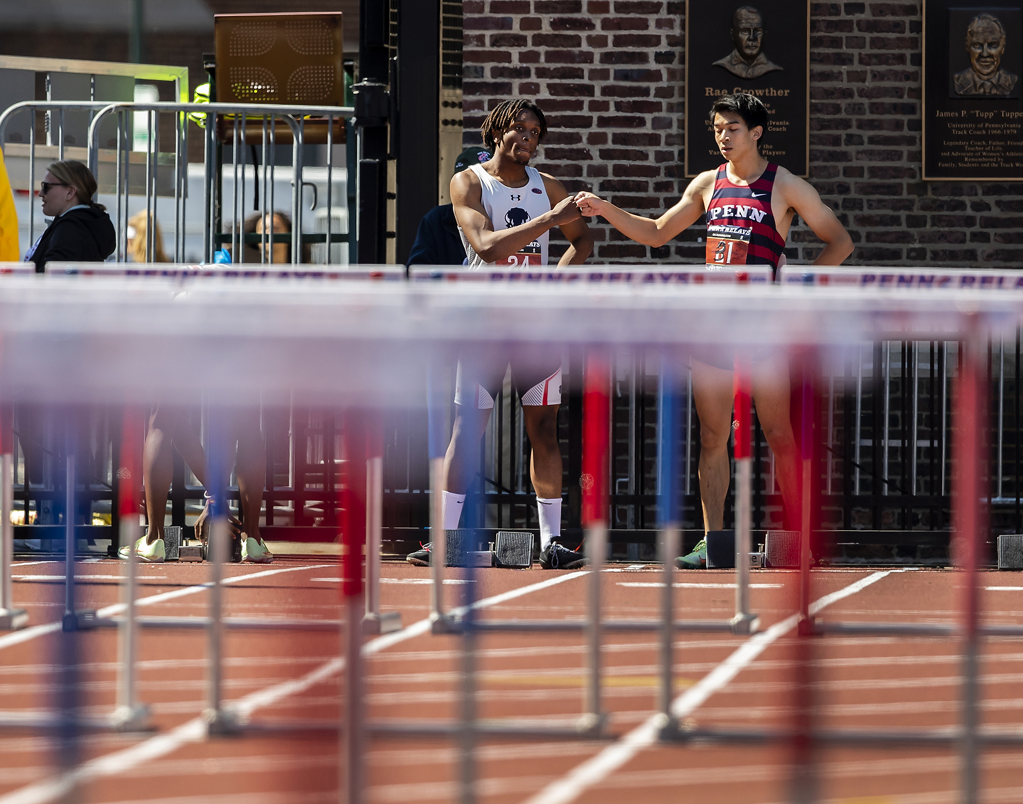 Two runners in front of hurdles on a track give each other a fist bump.