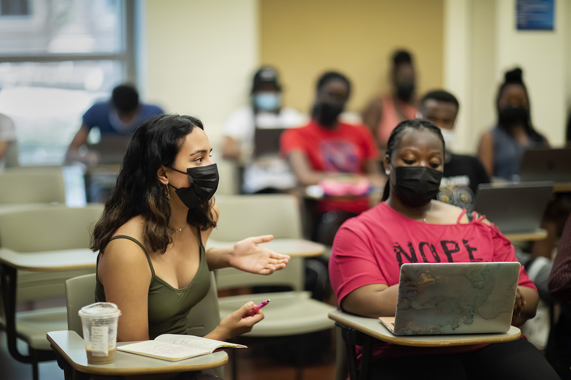Two masked students sit in the front of a classroom, one with a laptop, the other speaking