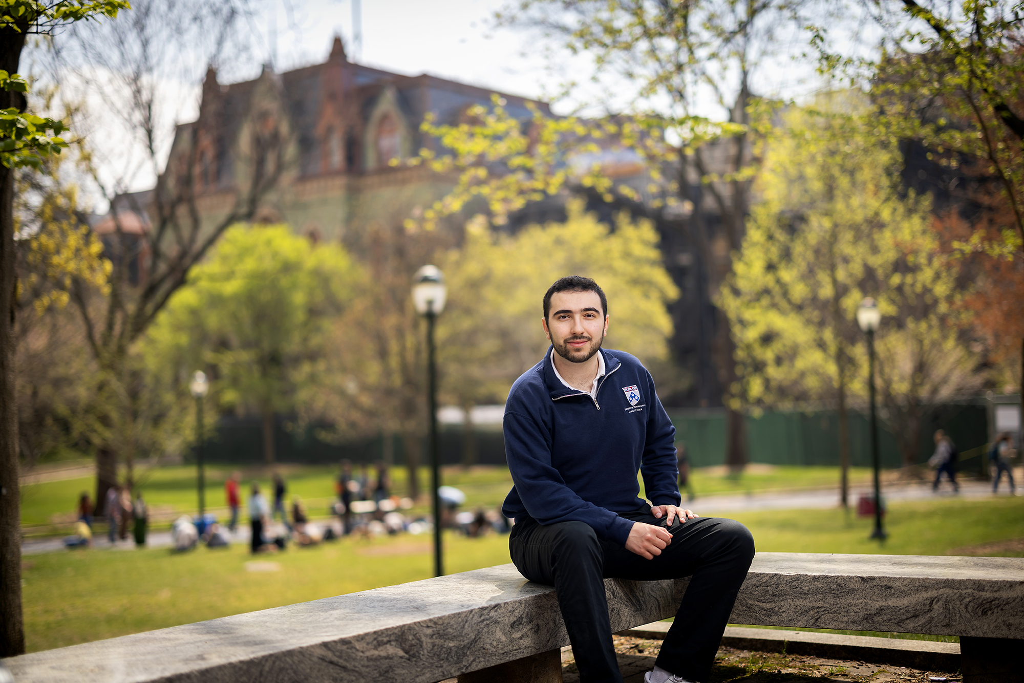 Ara Patvakanian sits on a low wall on Penn campus, with College Hall and the lawn full of students behind him.
