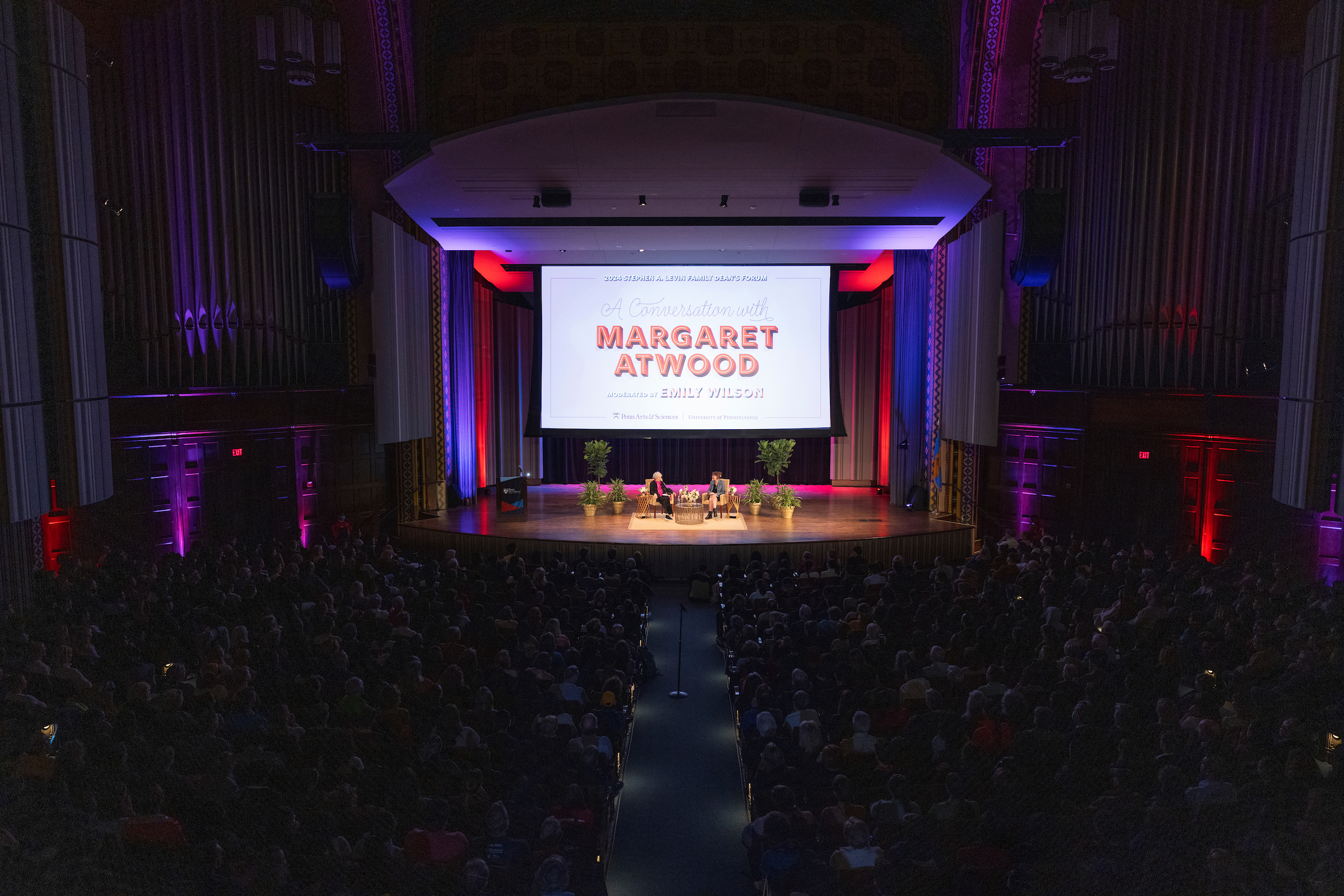 An auditorium with the lights down and the large screen lit that reads Margaret Atwood with Atwood and Emily Wilson seated on stage
