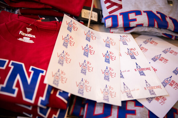 A collection of folded Penn T-shirts and sheets of "I voted" stickers in different languages are arranged on a table.