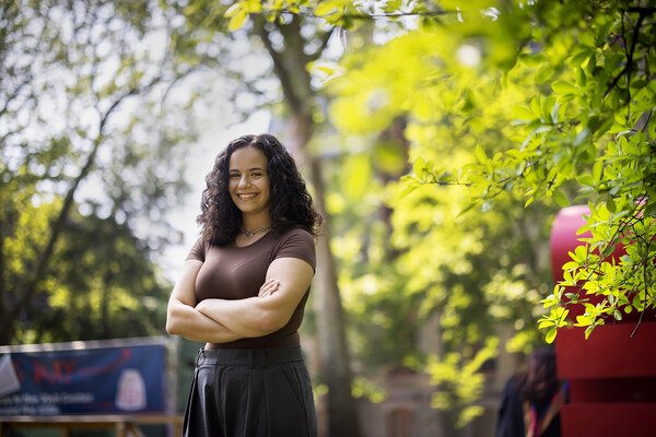 Amanda Yagerman poses with arms crossed, surrounded by trees with bright green leaves on Penn's campus.