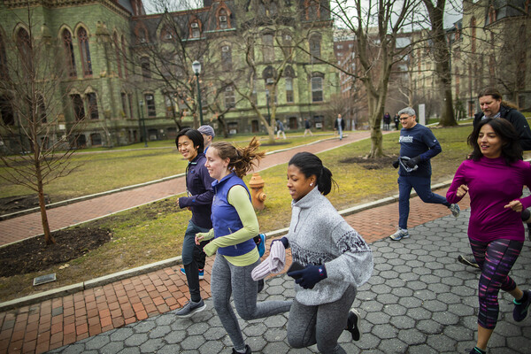 A group runs past trees and a green colored, Gothic-style building, Penn's College Hall.