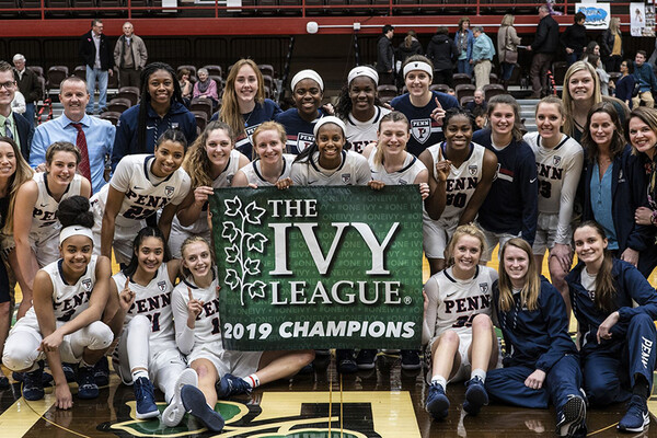 Members of the women's basketball team pose as a team with a banner that reads The Ivy League 2019 Champions on the court 