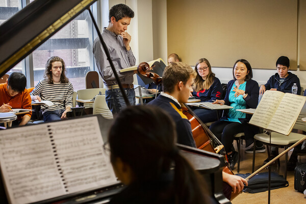 Student playing the cello in front of class and professors and another student playing the piano. 