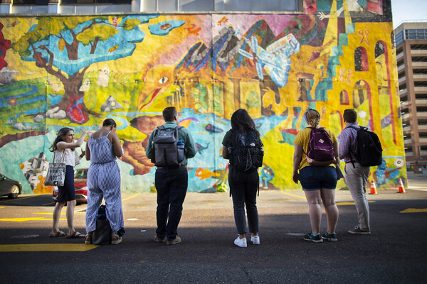 Six people standing in a parking lot looking at a colorful mural on the wall of a building. 