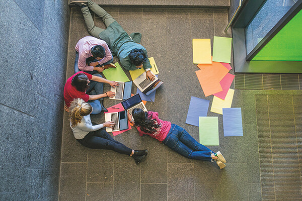 aerial view of five kids laying and sitting on the ground working on laptops in a school hallway