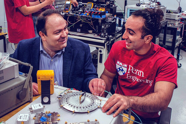 Firooz Aflatouni and a member of his lab sit at a table in his lab surrounded by engineering equipment.