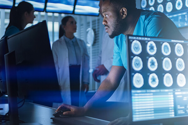 One person stands before two computer monitors while two people stand behind them, all in lab garments or white coats, one computer has brain scans on the screen