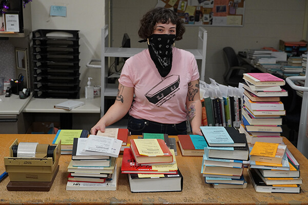 A person wearing a face covering stands in front of a table of books prepared for pickup.