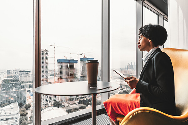 Professional African American person using a digital tablet looking out the windows on the top floor of a city office skyscraper.