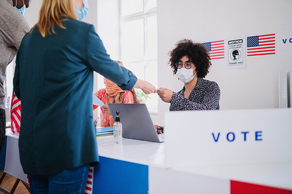 Person seated at a table with laptop wearing a face mask is taking an ID from a person standing before them, a polling place with a VOTE sign and American Flag pictures on the wall.
