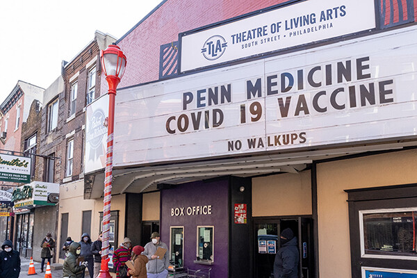 Marquee outside the Theatre of Living Arts that reads PENN MEDICINE COVID-19 VACCINES NO WALKUPS with masked people standing on sidewalk outside.