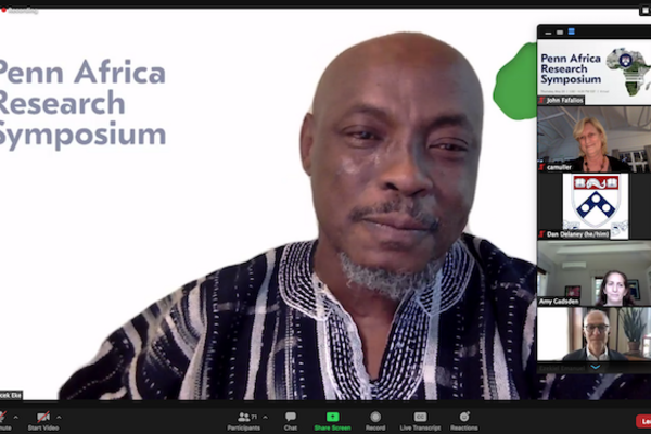 A Zoom video event shows man in a traditional African print shirt with the words "Penn Africa Research Symposium" on the background behind him, as six participants are seen in video images on the right side of the screen, stacked vertically.