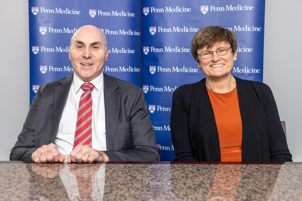 Two people seated at a table in front of a panel that reads "Penn Medicine" many times over. The person on the left is wearing a gray suit, with a white shirt and red tie. The person on the right is wearing glasses, an orange shirt, and a black cardigan.