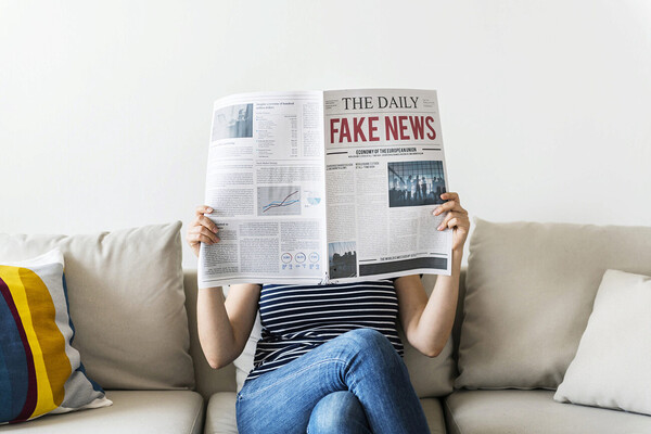 Person sitting on a couch reading a newspaper in front of their face, headline reads FAKE NEWS.